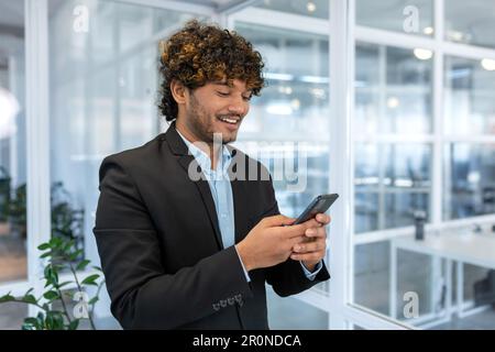 Successful financier investor works inside office at work, businessman in business suit uses telephone near window, man smiles and reads good news online from smartphone. Stock Photo