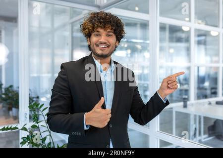 Smiling businessman inside office pointing fingers to direction, happy hispanic boss in shirt Stock Photo