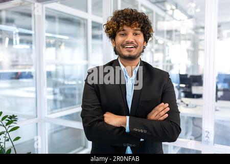 Portrait of successful hispanic boss, businessman in business suit looking at camera and smiling, man with crossed arms working inside modern office building. Stock Photo