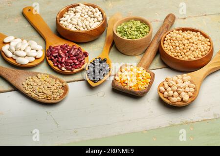 Beans, lentils, chickpeas, soybeans and corn kernels in wooden spoons and bowls on a green wooden table Stock Photo