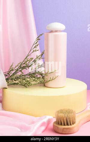 A pink bottle of moisturizing facial lotion sits on a yellow podium against a pink and lilac background.  Next to a wooden brush for massage Stock Photo