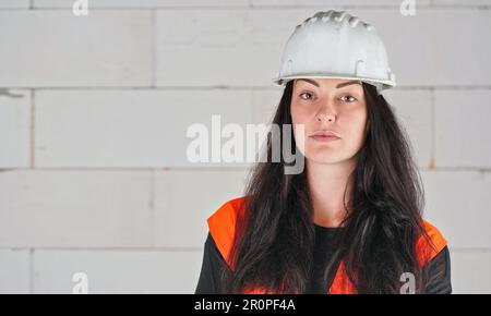 Young woman in white hard hat and orange high visibility vest, long dark hair, hands crossed, looking into camera. Blurred construction site wall back Stock Photo