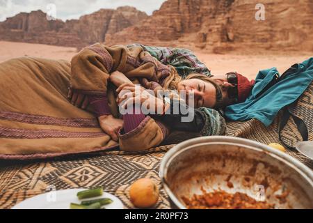 Man and younger woman wearing traditional Bedouin warm coat - bisht -  resting, lying down on ground blanket near off road vehicle back, blurred Wadi Stock Photo