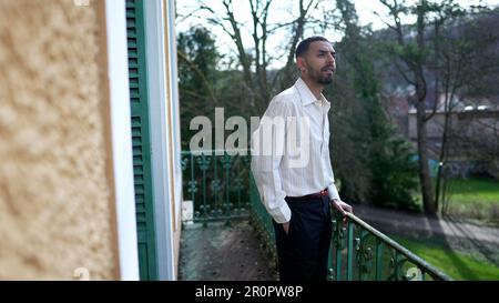 One contemplative young Middle Eastern man stands outside in home balcony with thoughtful expression Stock Photo