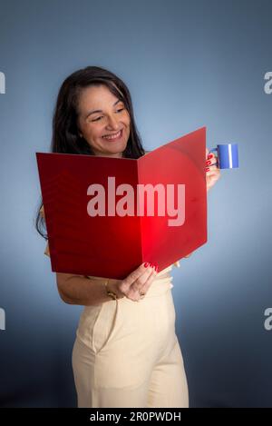 A young professional woman looking through a red folder while enjoying a cup of coffee in her office Stock Photo