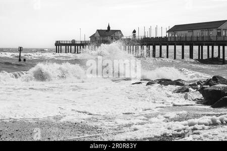 A wave crashes against rocks on the beach in Southwold, Suffolk, in front of the old pier. The image is black and white. Stock Photo