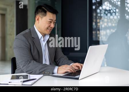 Successful mature asian working inside office using laptop, man typing on keyboard and smiling, businessman satisfied with work and achievement results, programmer at work. Stock Photo