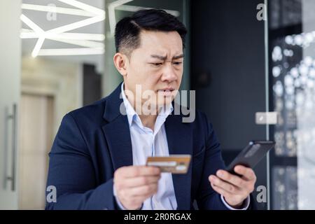 Cheated online buyer, asian businessman in office close up trying to make bank transaction and purchase in online store, man unhappy holding bank credit card and phone Stock Photo
