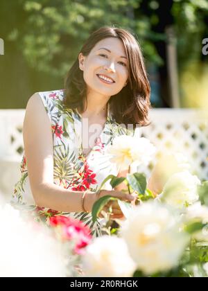 Wide smiling woman is admiring of blooming roses in public park. Summer vibes. Tropical plants and flowers in bloom in garden. Stock Photo