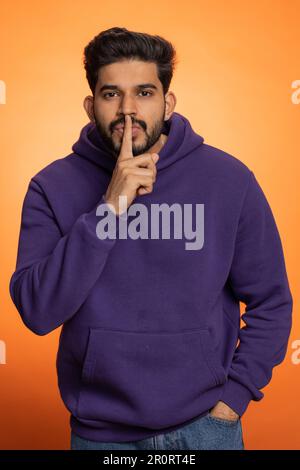 Shh be quiet please. Young indian man presses index finger to lips makes silence gesture sign do not tells secret, stop gossip. Handsome bearded hindu guy isolated alone on orange studio background Stock Photo