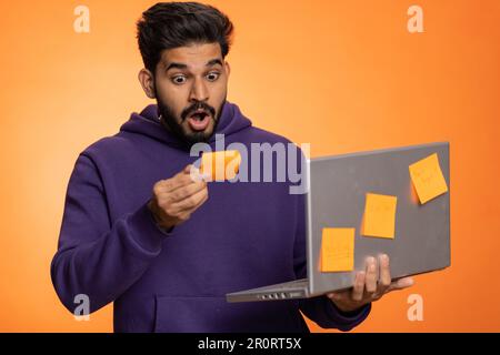 Exhausted indian freelancer man with pasted sticker notes use laptop computer having concentration problem creative crisis. Nervous hindu guy confused by big routine work loading on orange background Stock Photo