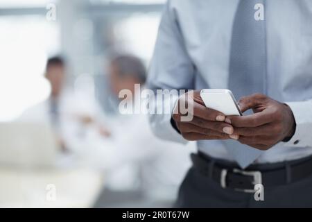 The world of business at your fingertips. A close-up of an African businessmans hands holding a cellphone with colleagues working in the background. Stock Photo