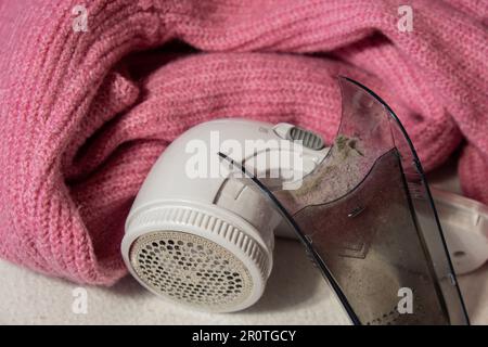 Anti-pilling razor. Device for shaving clothes. Anti-Plush fabric Shaver. Electric portable sweater pill defuzzer Lint remover from pink acrylic or wool sweater. Electric device fabric shaver after cleaning and collected fluff lint. Closeup Pilled sweater. Old used clothes with pilling.  Stock Photo