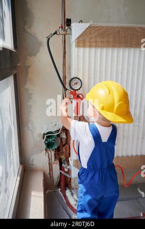 Kid using air compressor manometer while checking gas tightness of radiator in apartment under renovation. Child filling pipes with pressurized air to inspect for leaks in heating system. Stock Photo