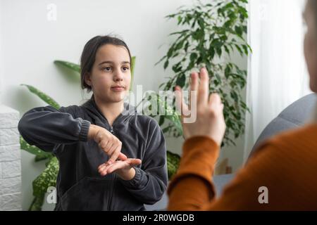 daughter talk with middle aged mother people using sign language, family sitting on armchair side view, teacher teach teenager deaf-mute girl to Stock Photo