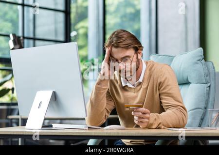 Cheated and disappointed businessman working inside office, man holding bank credit card and trying to make a purchase in online store, online fraud and deception Stock Photo