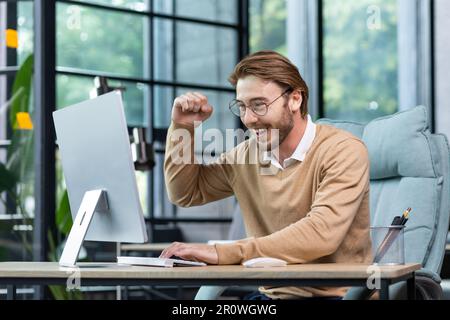 A young man sits in the office, looks at the laptop monitor and rejoices. Screams with happiness, makes victorious hand gestures. Received good news, successful deal, project, playing online games. Stock Photo