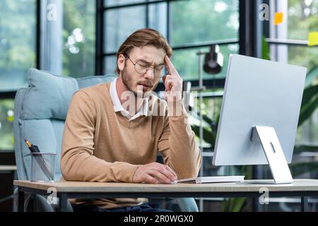 Overtime, deadline. Tired young male programmer, freelancer sits in the office and tries to work at the laptop. Bored, he sits at the table and rests his head on his hand. Stock Photo