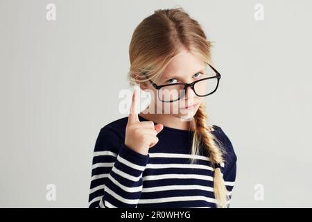 Strict, serious and portrait of a child with a gesture isolated on a white background in a studio. Rude, smart and a young girl wearing glasses and Stock Photo