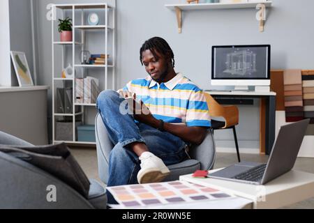 African American designer working online on tablet pc while sitting on armchair in office Stock Photo