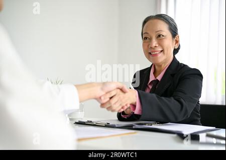 Smiling and happy senior Asian businesswoman or HR manager shaking hands with a candidate after a job interview. Stock Photo