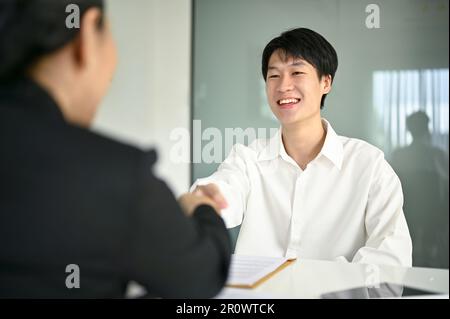 Smiling and confident young Asian male candidate shaking hands with a businesswoman after a job interview. Stock Photo