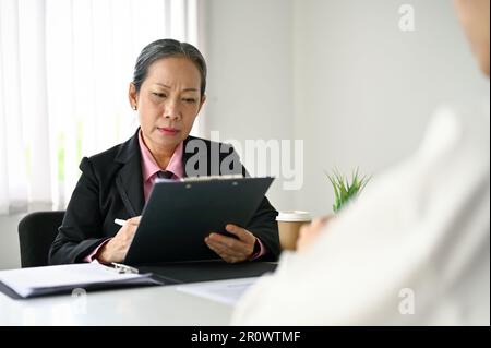 Professional and focused senior Asian female HR manager or recruiter reading a candidate's resume during a job interview. Stock Photo