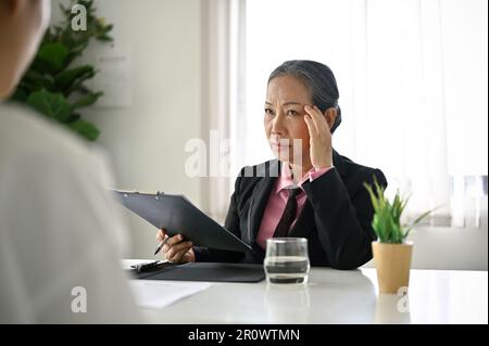 Stressed and upset senior Asian businesswoman or female recruiter listens to candidate answers during a job interview. Stock Photo