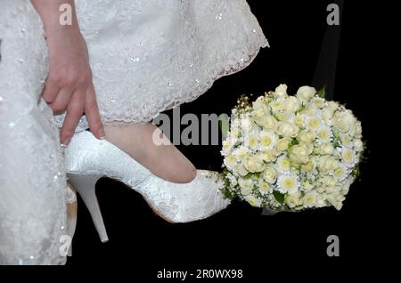 close-up of the bottom of a wedding dress and the hand of a woman touching a white wedding shoe, with a bouquet of small white flowers on a black back Stock Photo