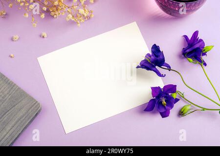 Blank card, notepad, glass of water and blue Aquilegia Alpina flowers on purple table. Top view, flat lay, mockup. Stock Photo