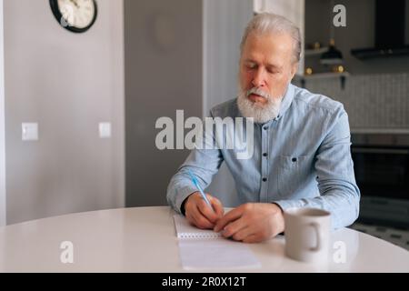 Portrait of focused grey haired senior older man writing making notes in organizer book, keep personal diary sitting at desk. Thoughtful mature male Stock Photo