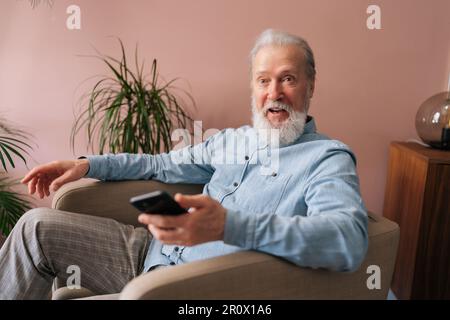 Portrait of happy excited gray-haired mature male holding smartphone using mobile online app, shocked looking at camera with open mouth sitting on Stock Photo
