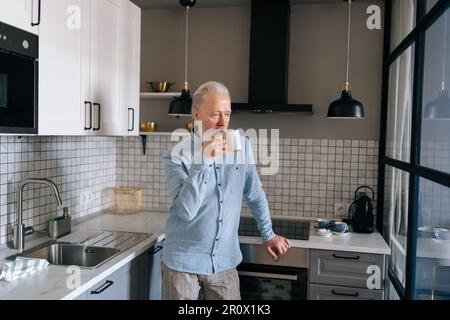 Medium shot of thoughtful gray-haired mature adult male drinking hot coffee and looking through window in morning at home. Elderly bearded man Stock Photo