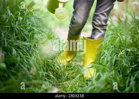 Boy walking with his wellies and a watering can in a grassy meadow. Conceptual image that can represent Earth Day and Environment Day. Stock Photo