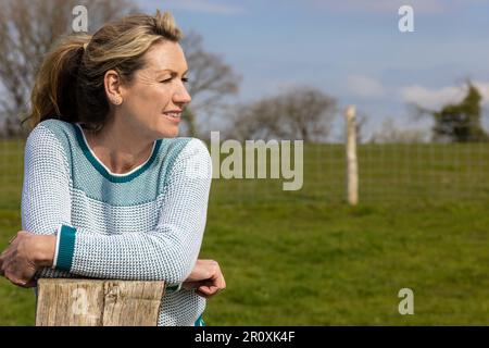 Attractive thoughtful middle aged woman leaning resting on a wooden fence post in the countryside or her garden Stock Photo