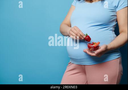 Midsection of a pregnant woman in blue t-shirt, holding glass bowl with sweet strawberries, isolated on blue background Stock Photo
