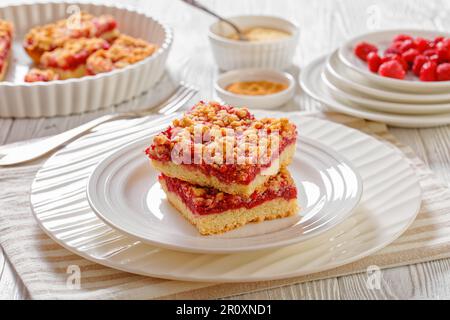 Raspberry Crumble Bars on white plate on white wood table with ingredients at background, horizontal view from above, close-up Stock Photo