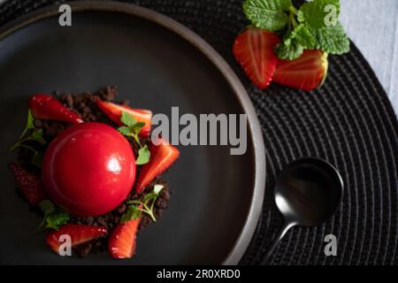 Selective focus of a sphere curd cake with strawberries and brownie. Top view. Dessert with smooth surfaces and mirror glaze. Black plate and spoon. Stock Photo