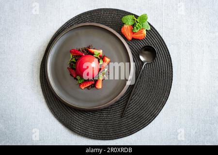Top view of a sphere curd cake with strawberries and brownie crumbs. Red dessert with smooth surfaces and mirror glaze. Black plate and spoon. Stock Photo