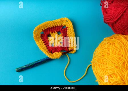 Granny square in yellow and red being crocheted, with two balls of yarn in yellow and red partly visible, all on an aqua blue background. Stock Photo