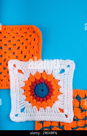 White Granny Square with Central Orange, Red and Blue Pattern. Two Orange Granny Squares behind all on a Kingfisher Blue Backgraound. Stock Photo