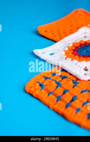 Three Crocheted Granny Squares, Two Orange, One White with Orange, Red and Blue Central Pattern, all on an Aqua Blue Background. Copy Space  to Left. Stock Photo