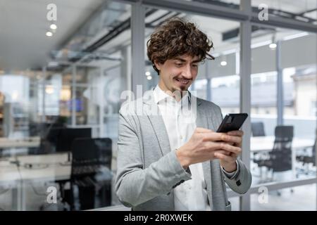 Successful businessman with beard standing near window, man using phone, happy boss typing message and browsing online pages on smartphone app inside office Stock Photo