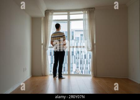 Young man standing in an empty room. New home buyer looking through a window out at the street in Brampton, Ontario Canada. Stock Photo