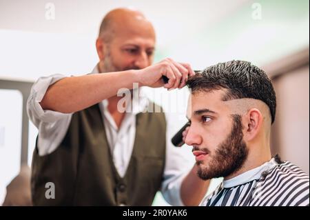 Focused male barber trimming hair of serious bearded client with comb and trimmer in barbershop Stock Photo