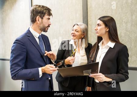 Group of coworkers in formal clothes standing together in modern office and talking about business strategy while looking at each other during meeting Stock Photo