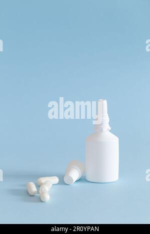 A bottle of nasal spray with the cap removed and some medication capsules placed next to it. White products on blue background. Vertical. Stock Photo