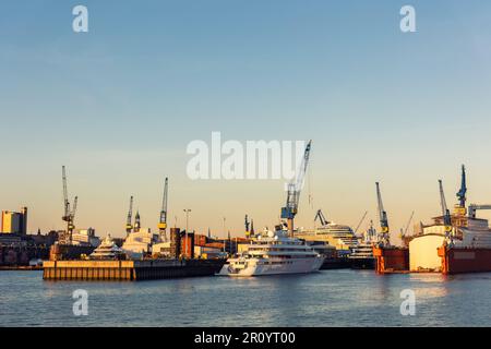 Scenic panoramic view large modern luxury super yacht at maintenance service against construction cargo cranes at dry dock shipyard in Hamburg port Stock Photo