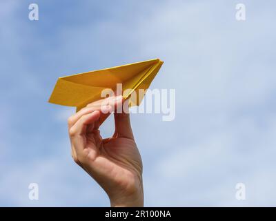 A hand holding a yellow paper airplane against a cloudy sky. Close up. Stock Photo