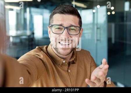 Close-up photo. Young man freelancer, programmer takes a selfie on the phone and talks on a video call in the office. He holds a device in his hand, smiles at the camera. Stock Photo
